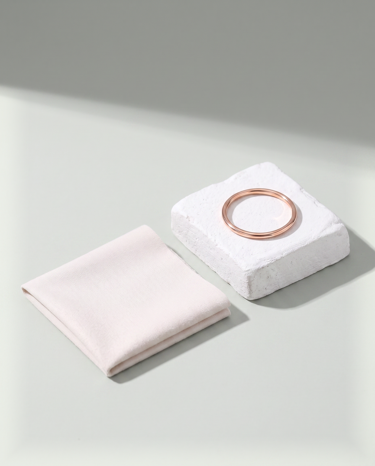 A folded soft polishing cloth beside a slender rose gold band resting on a white plaster block