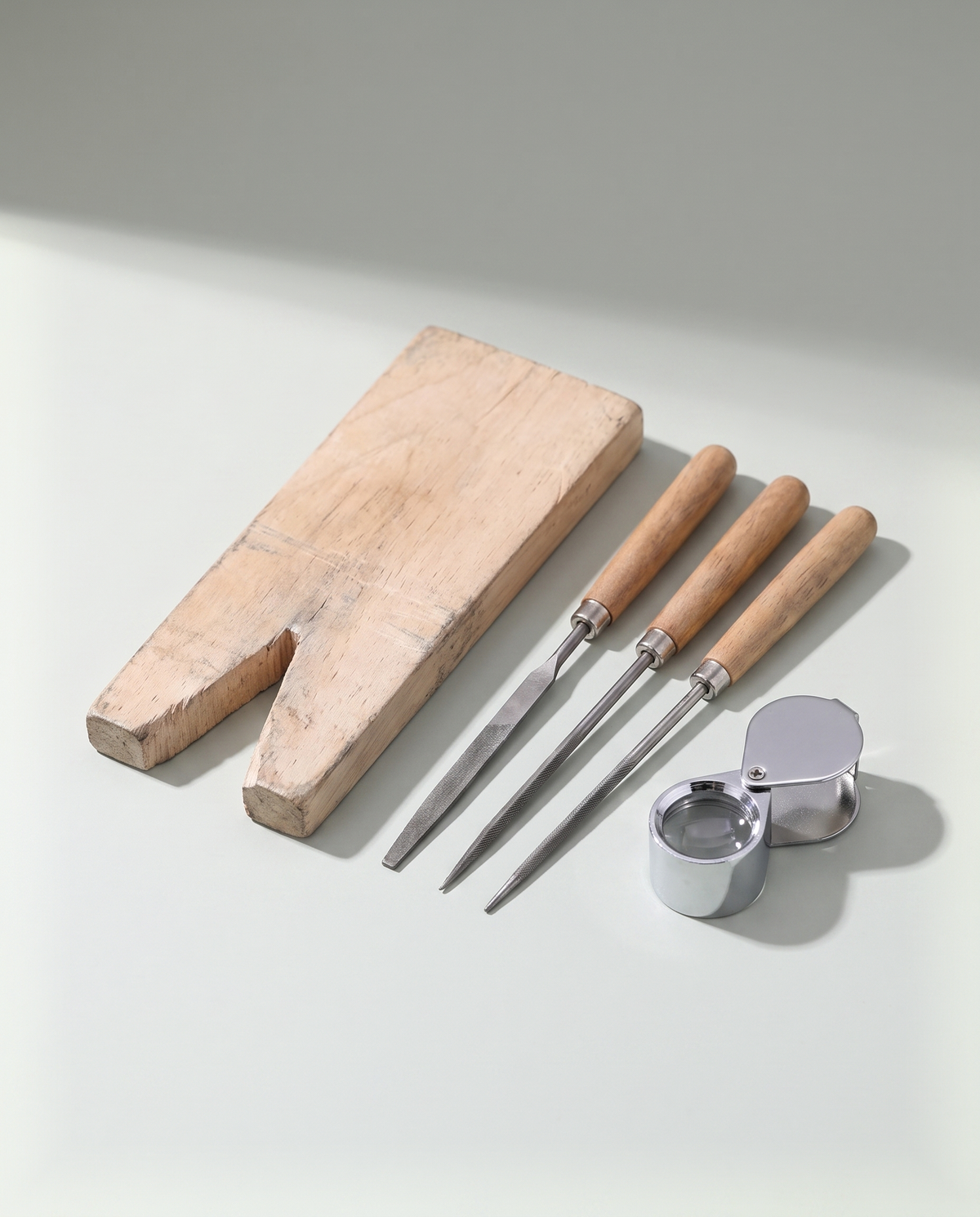 A jeweller's wooden bench pin laid beside three fine steel needle files and a folding loupe