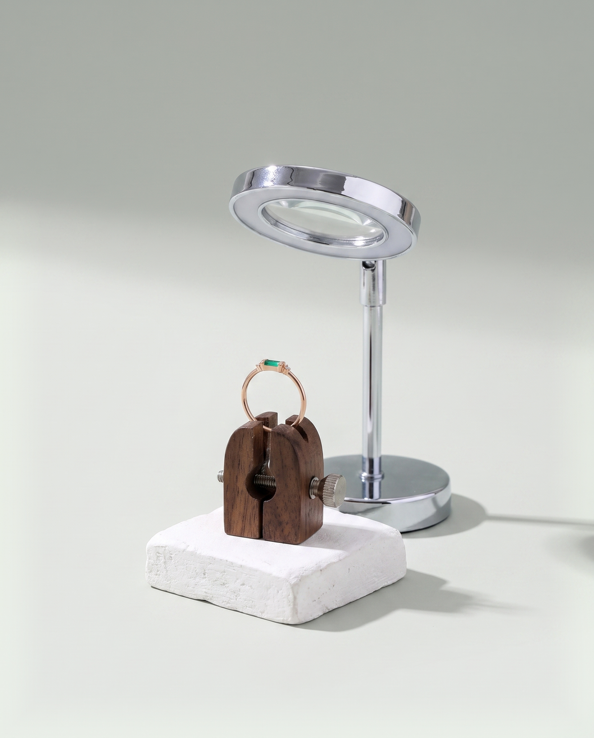 A rose gold ring held upright in a wooden setting clamp beneath a bench magnifier