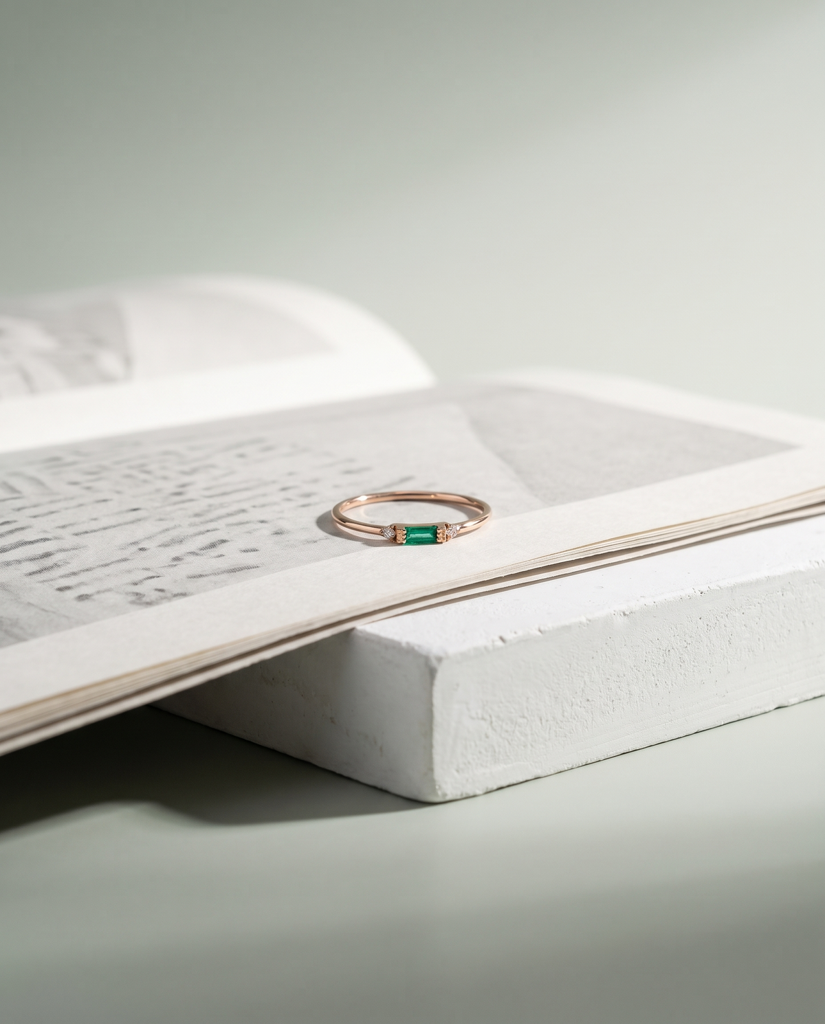 A rose gold ring with an emerald baguette resting on an open printed page, on the bench plinth