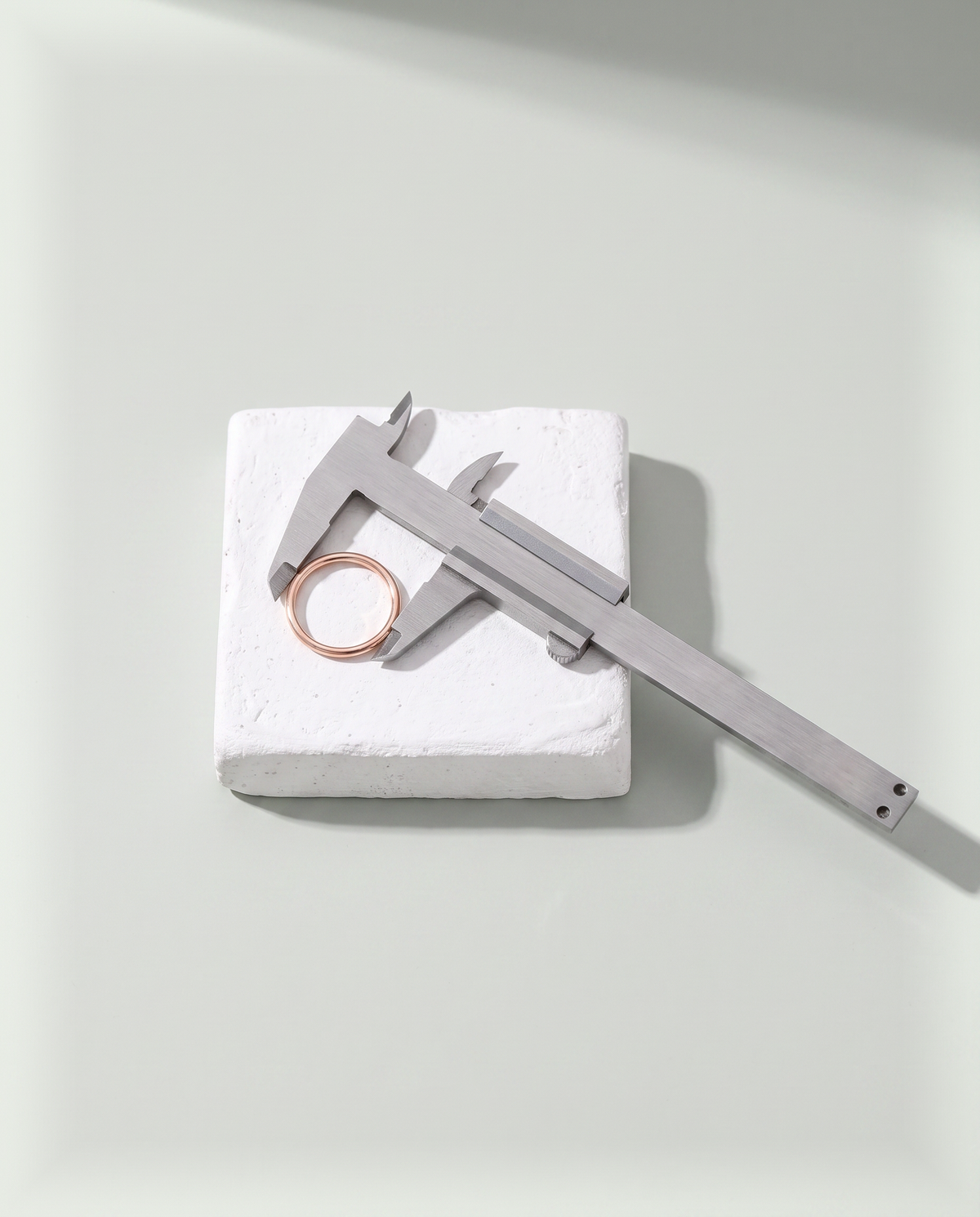 A pair of steel calipers spanning the inner width of a plain rose gold band on a white plaster block